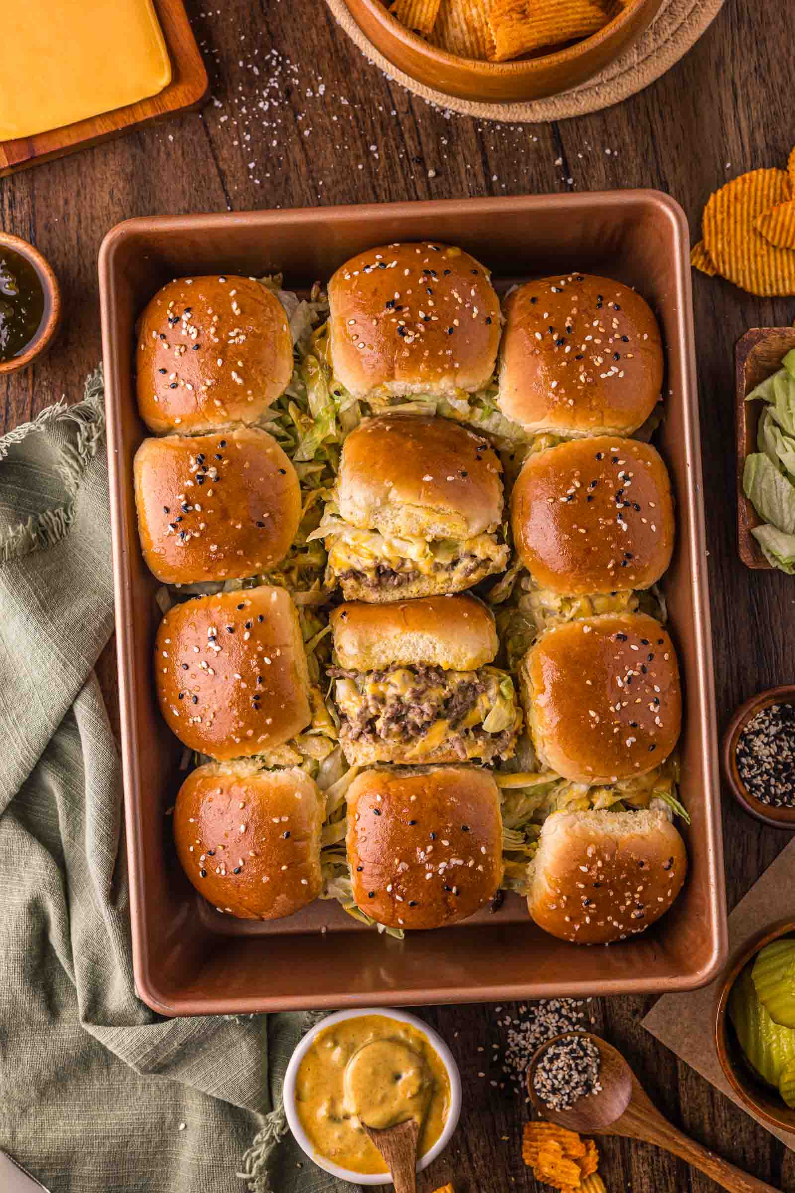 A baking dish filled with cheeseburger sliders on sesame seed buns, with shredded lettuce and cheese, surrounded by condiments and potato chips on a wooden table.