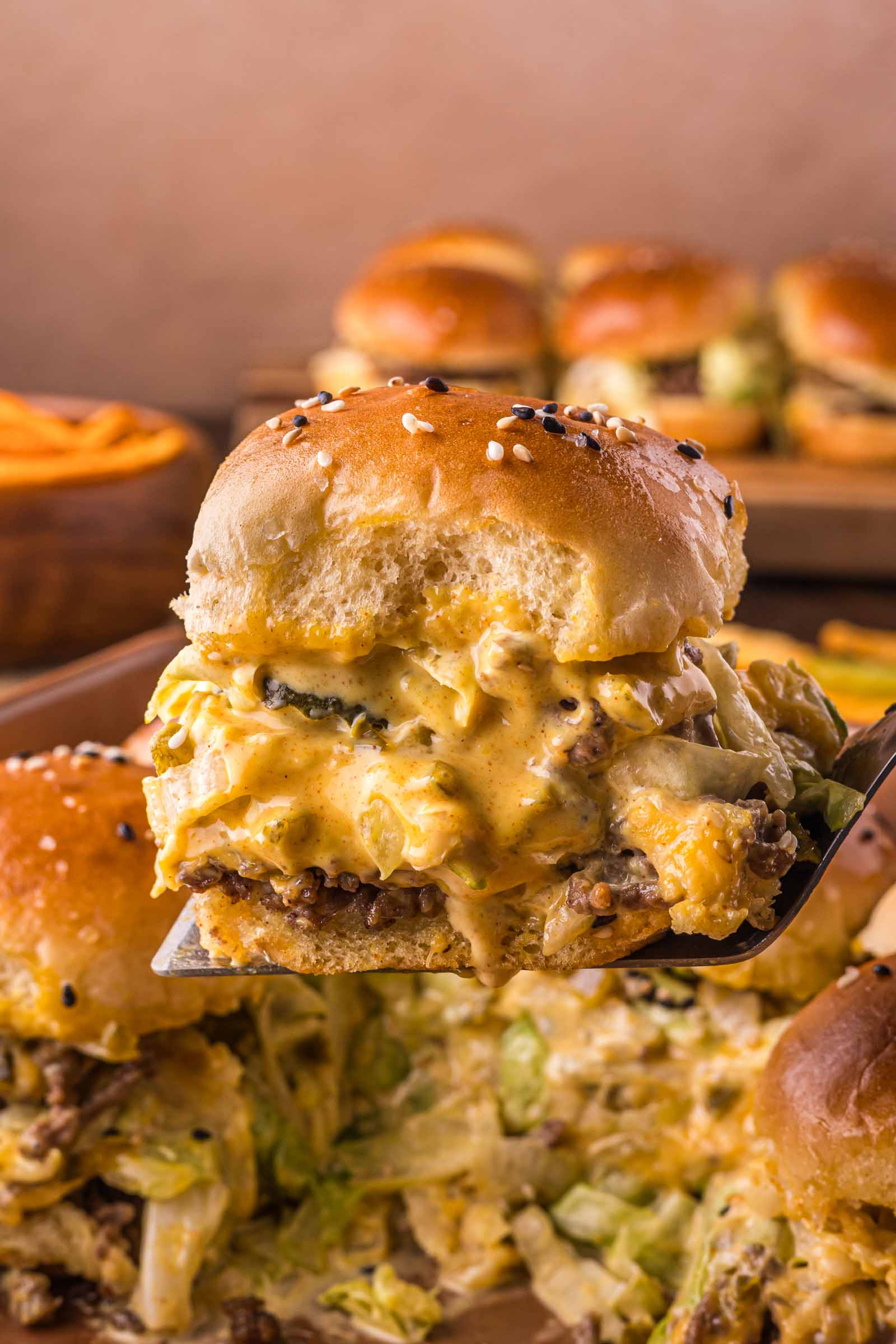 A close-up of a cheesy big mac slider with layers of ground beef, melted cheese, and pickles on a sesame seed bun, being lifted from a tray. More sliders and fries are in the background.