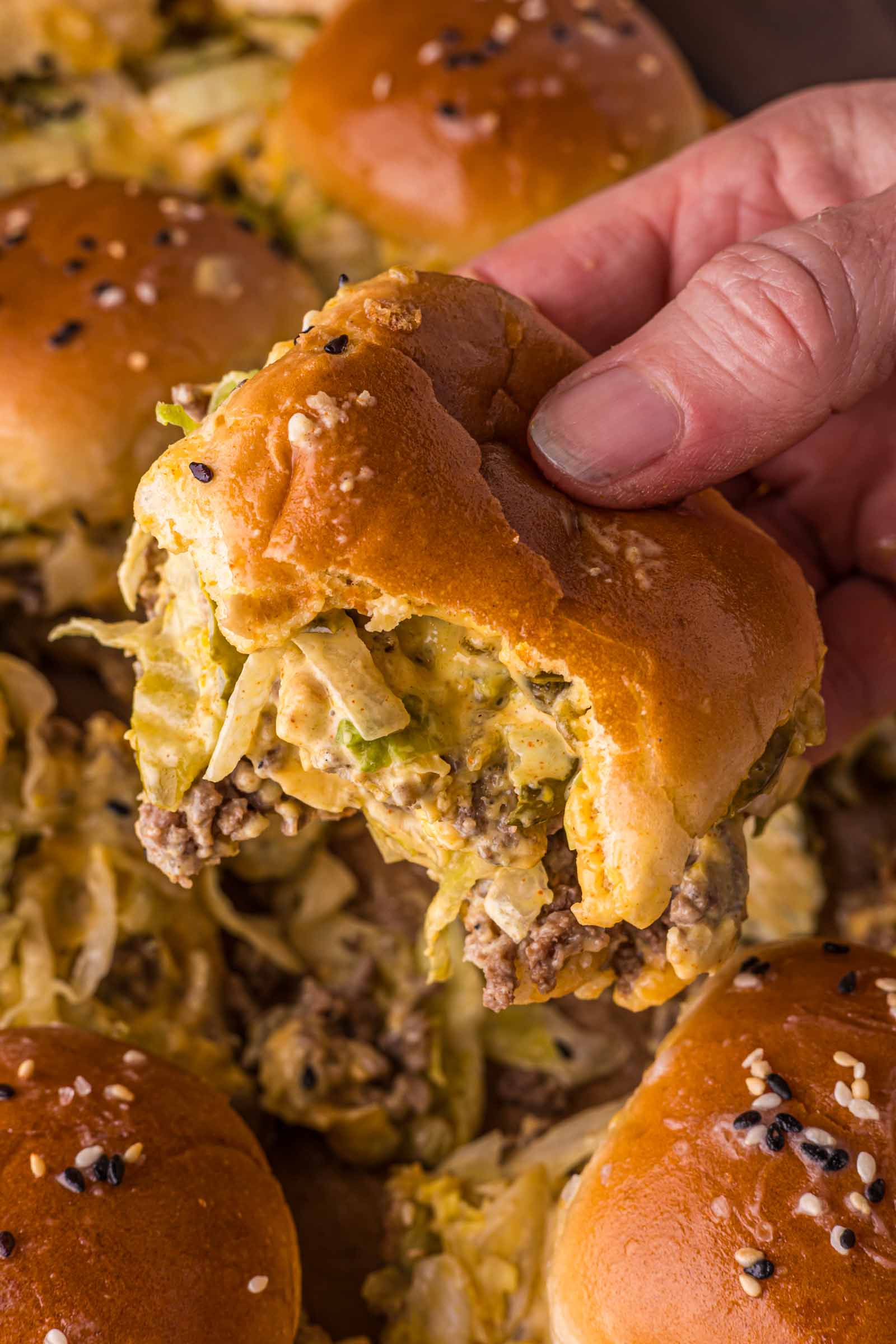 A hand holding a partially eaten big mac slider with ground beef, melted cheese, and shredded lettuce in a golden bun topped with sesame seeds.
