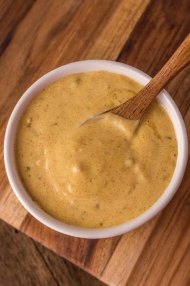 A small white bowl filled with creamy, orange-colored sauce with visible herbs, stirred with a wooden spoon on a wooden surface.