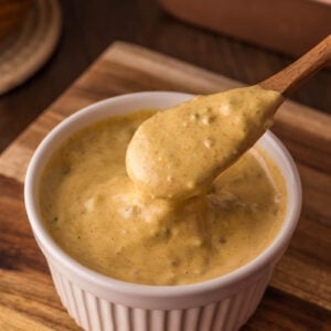 A wooden spoon scoops thick, creamy big mac special sauce with specks from a white ramekin on a wooden surface.