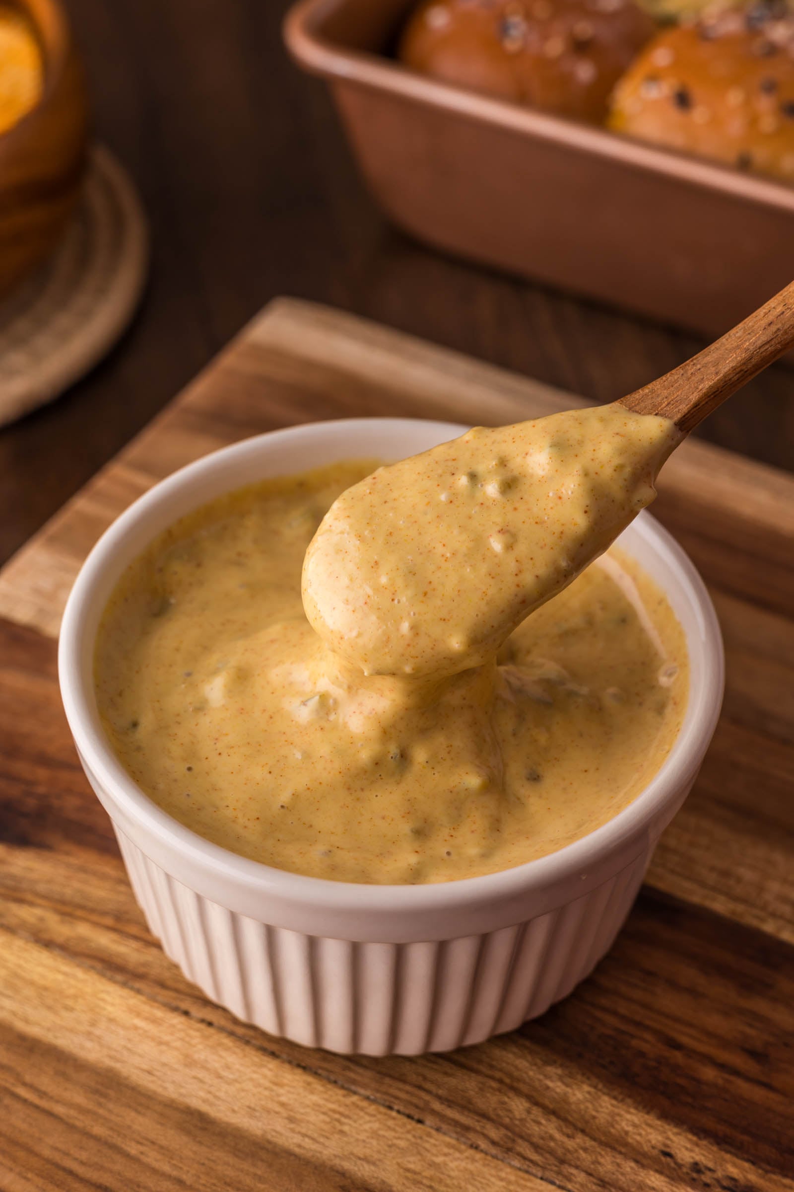 A wooden spoon scoops thick, creamy big mac special sauce with specks from a white ramekin on a wooden surface.