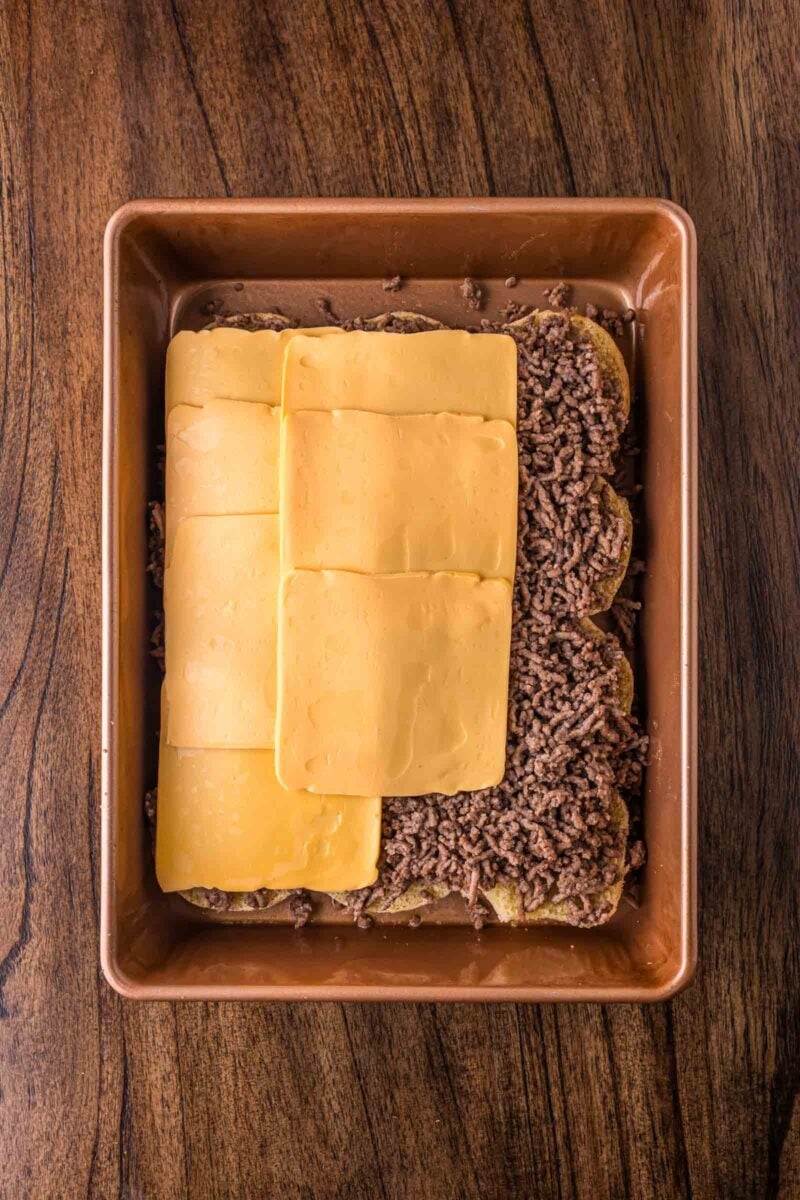 A baking dish with the bottom layer of sliders with cooked ground beef, sliced cheddar cheese, and what appears to be a bottom layer of tater tots on a wooden surface.