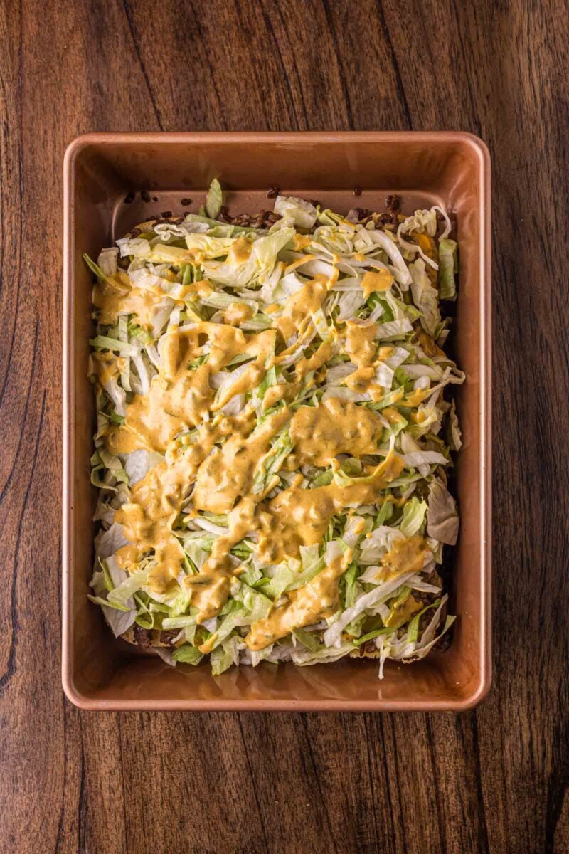 A rectangular baking dish filled with big mac sliders bring built - opened to show the layer of chopped lettuce and big mac sauce over the burger layer placed on a wooden surface.