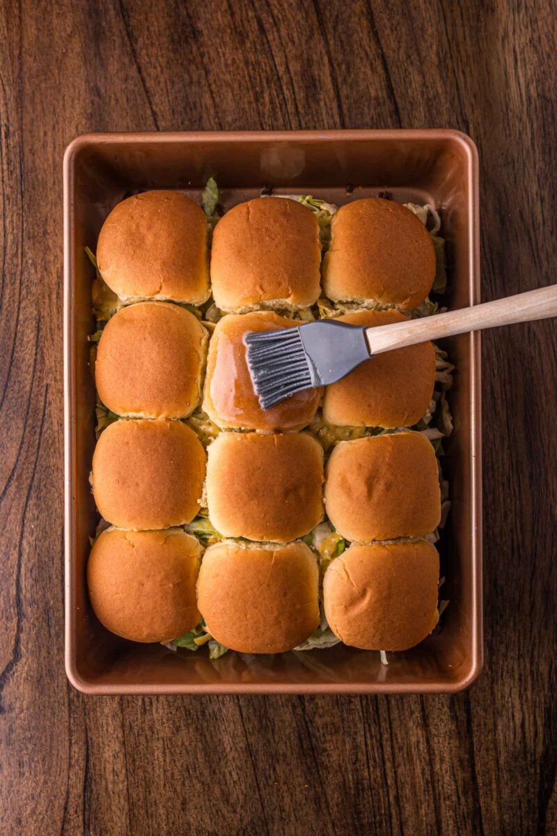 A baking dish of twelve sandwich sliders with golden buns, being brushed with a glaze using a pastry brush, on a wooden surface.