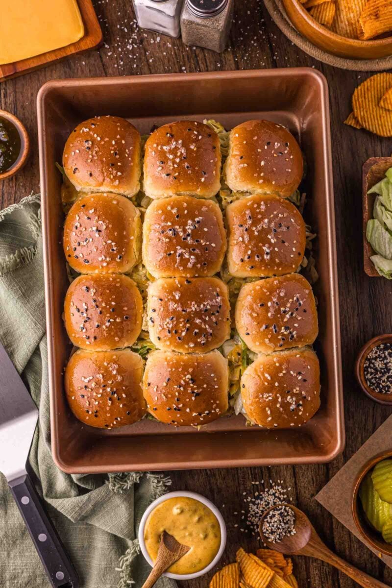 A baking dish filled with twelve golden-brown big mac sliders topped with sesame seeds, surrounded by condiments, chips, lettuce, and a knife on a wooden table.