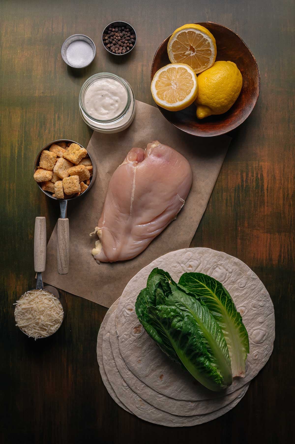 A raw chicken breast, romaine lettuce, tortillas, parmesan cheese, croutons, Caesar dressing, lemon halves, salt, and pepper are arranged on a tabletop.