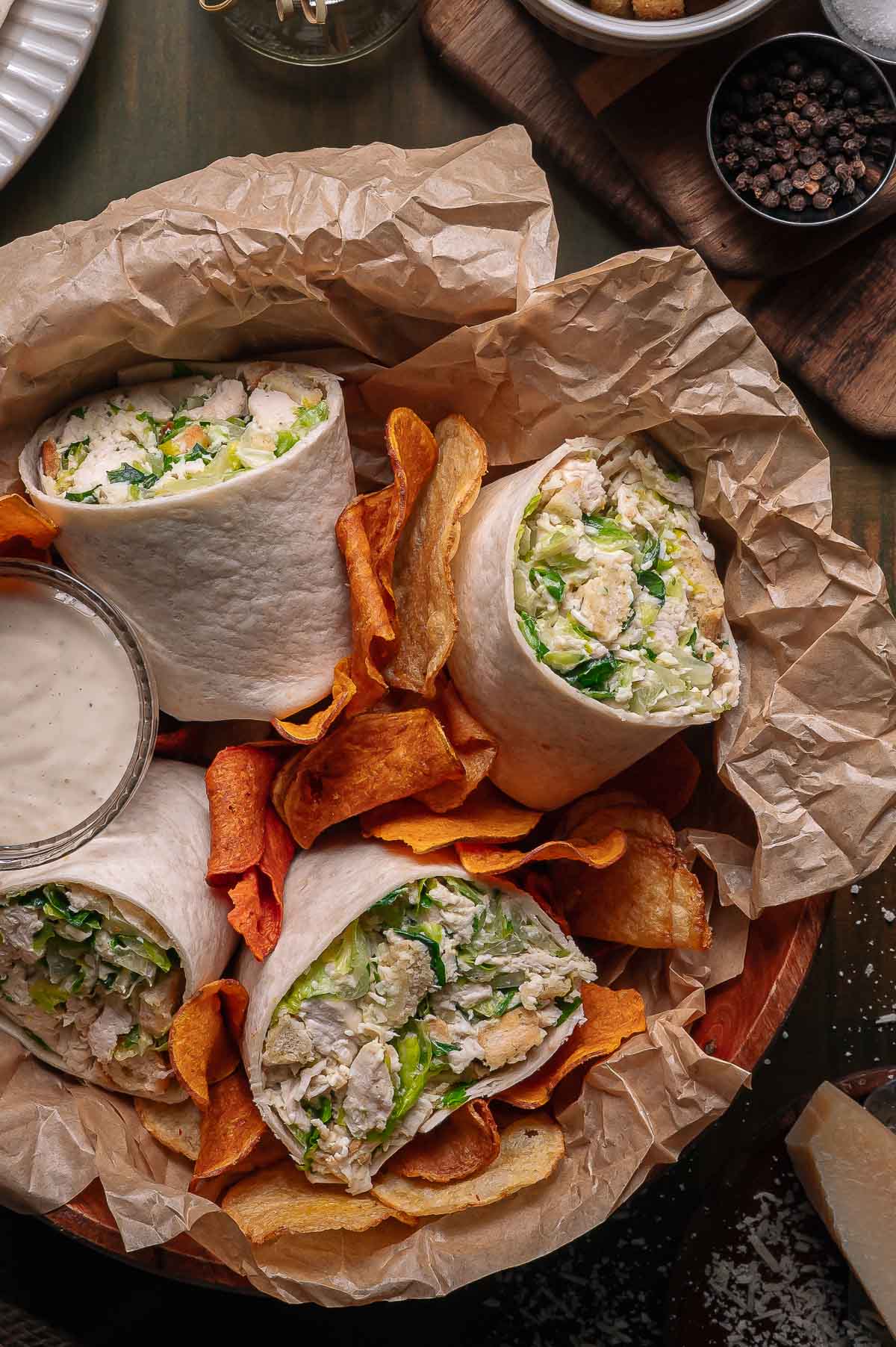 A basket lined with parchment paper holds halved chicken caesar wraps with lettuce, served with vegetable chips and a side of ranch dip.