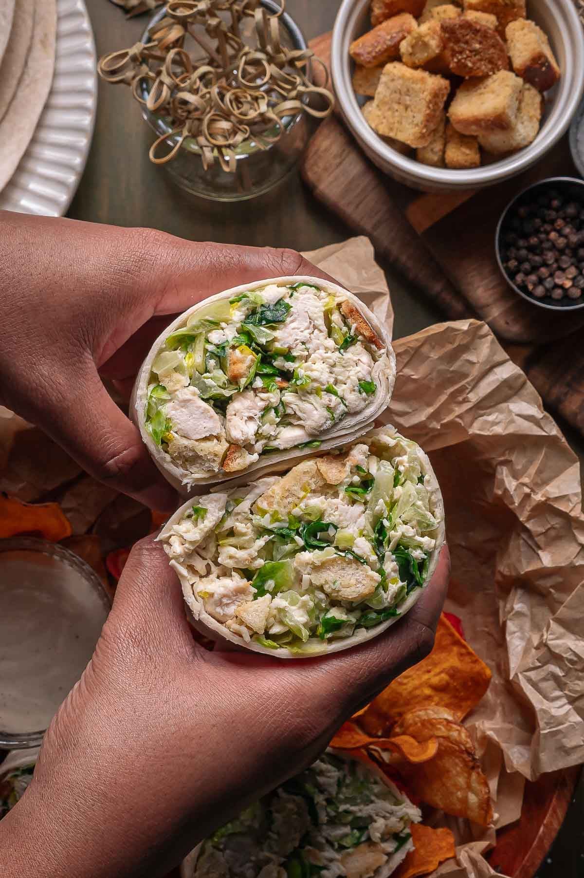 Two hands hold a halved wrap filled with chicken caesar salad with chips, croutons, and sauce visible on a wooden table.