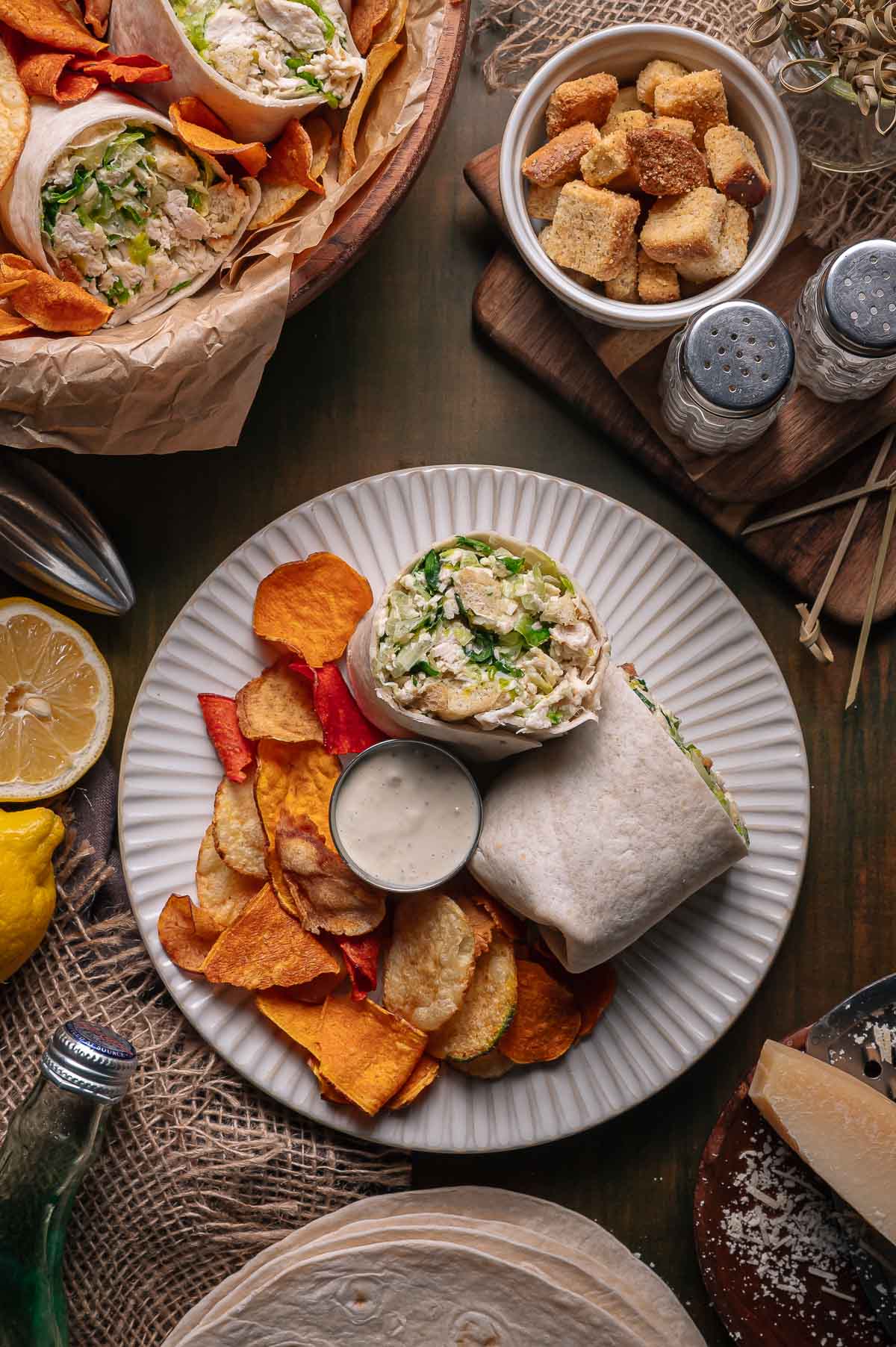 A plate with a chicken caesar wrap cut in half, vegetable chips, and a cup of dipping sauce. Nearby are a bowl of croutons, salt and pepper shakers, and fresh lemon halves.