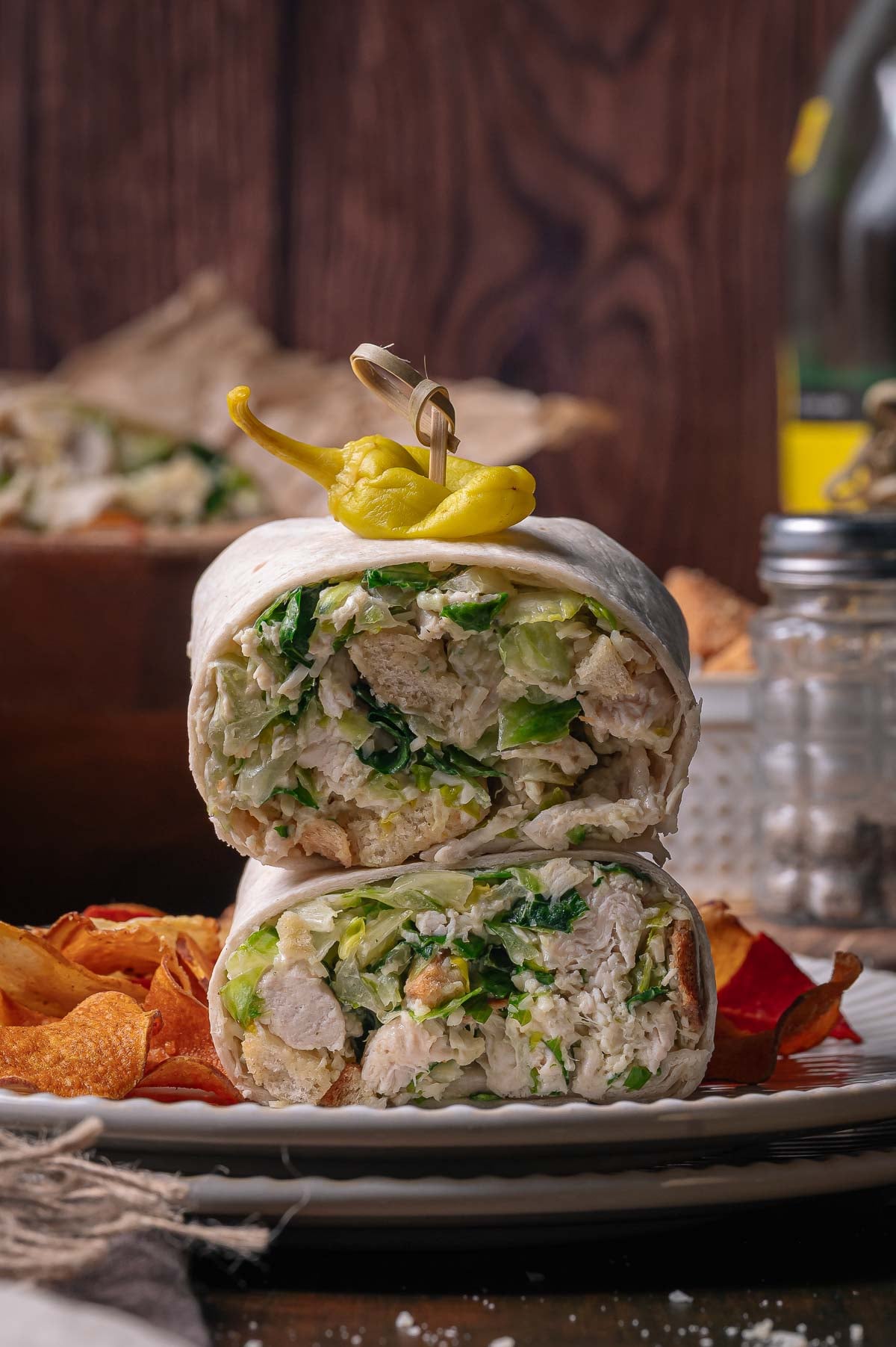 A chicken caesar salad wrap cut in half and stacked on a plate, topped with a yellow pepper, served with potato chips.