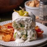 A sliced wrap filled with chicken caesar salad, topped with creamy dressing, served with potato chips and garnished with a pepper on a white plate.