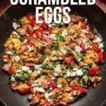 A skillet filled with Greek scrambled eggs, featuring tomatoes, spinach, feta, and sausage, with text overlay promoting a recipe from girlcarnivore.com.