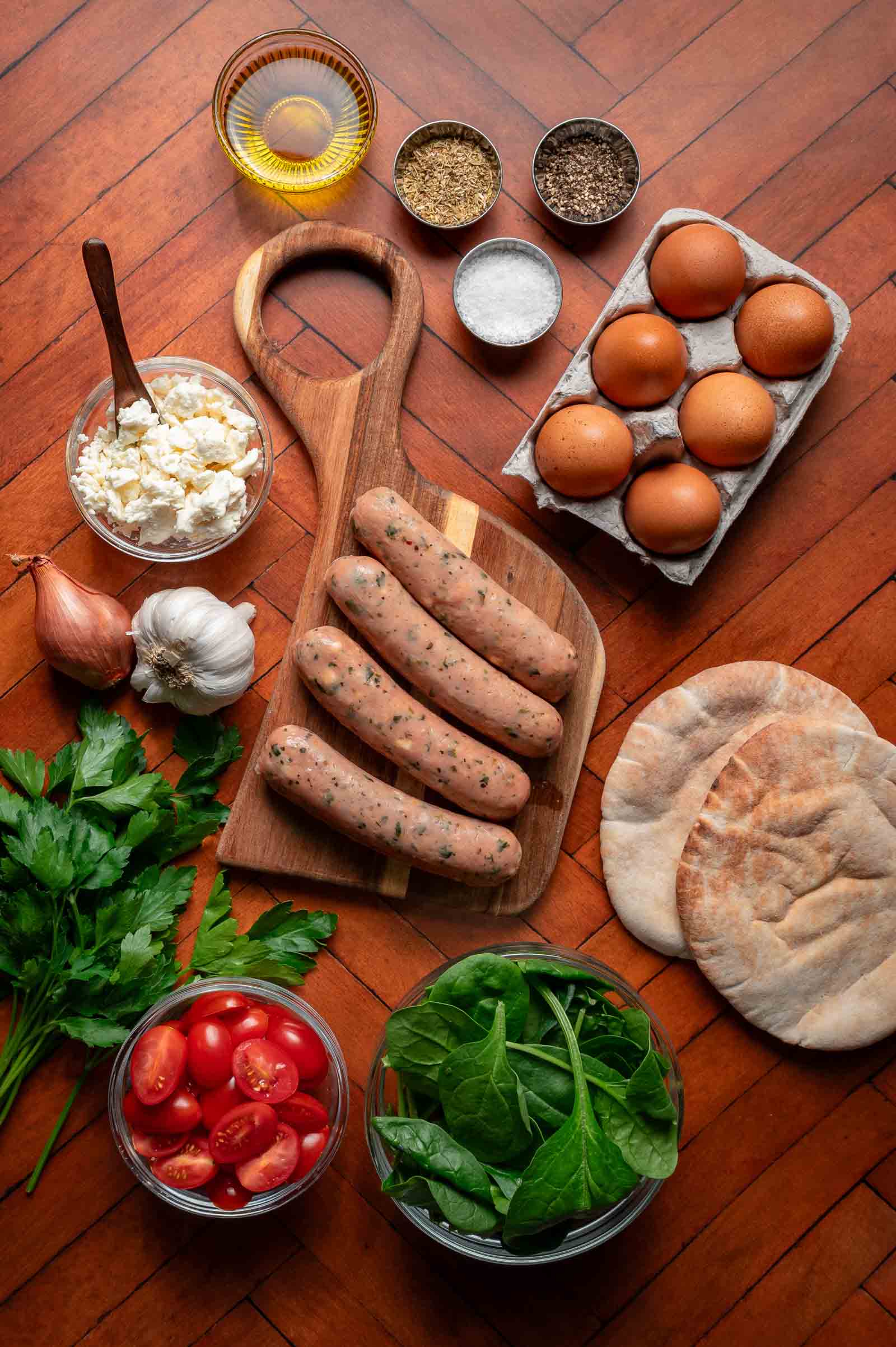 A wooden board with four uncooked sausages is surrounded by eggs, pita bread, spinach, cherry tomatoes, parsley, feta, a shallot, garlic, oil, salt, and pepper on a wooden surface.