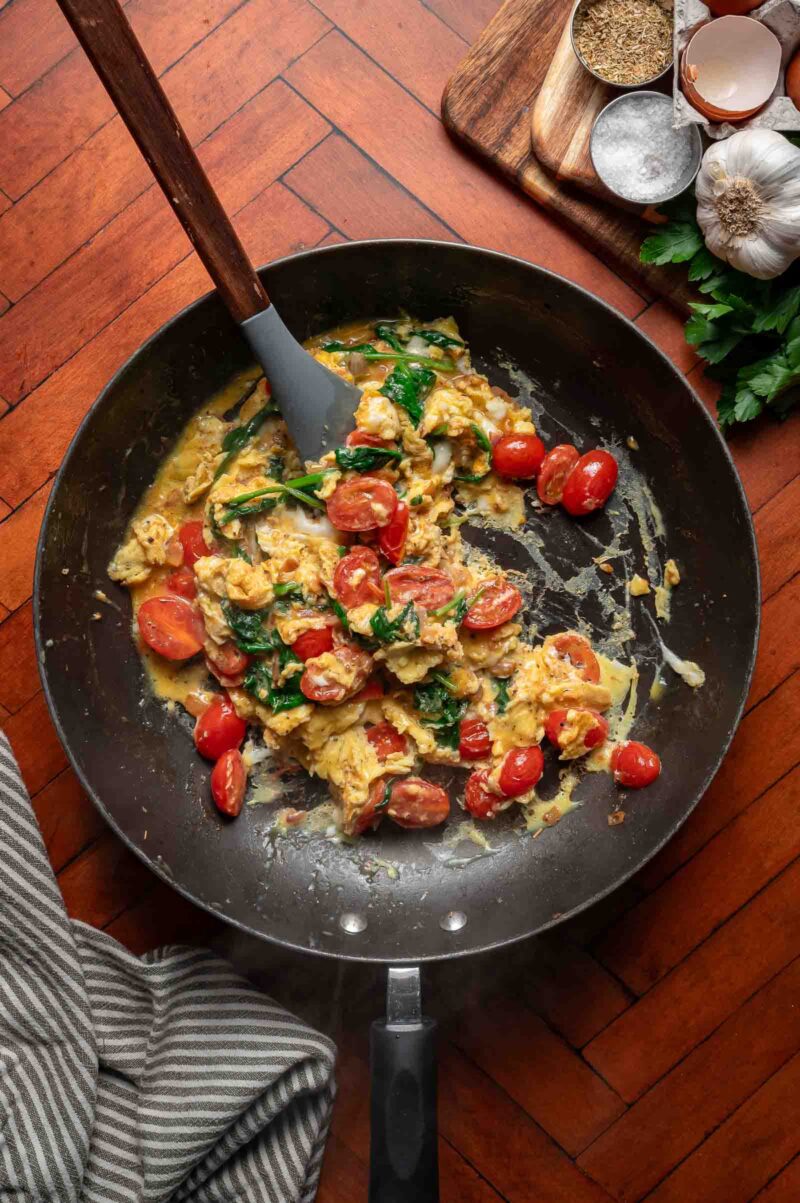 Greek scrambled eggs with cherry tomatoes and spinach in a black skillet on a wooden floor, with a spatula resting in the pan. Nearby are garlic, herbs, and spices on a wooden board.