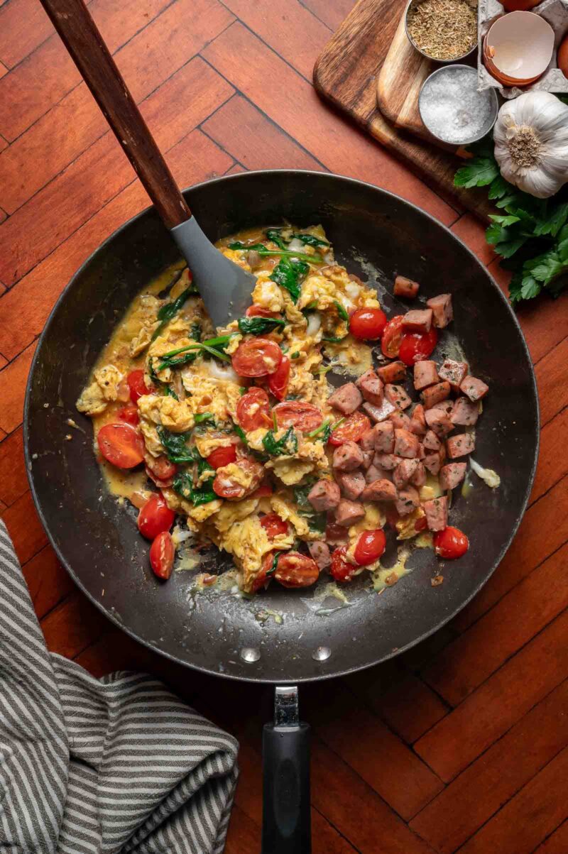 Greek scrambled eggs with spinach and cherry tomatoes next to diced sausage pieces cooking in a black skillet on a wood surface, with seasoning, parsley, and garlic nearby.