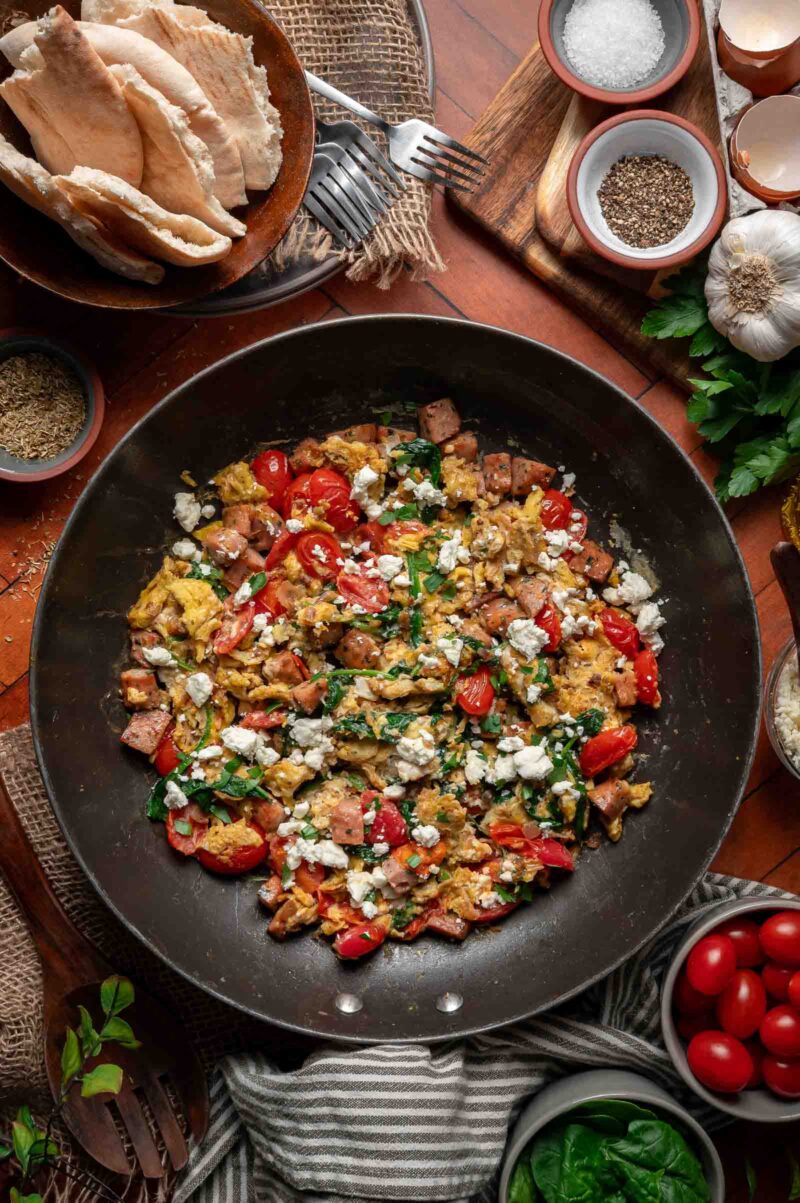 A skillet filled with Greek scrambled eggs, diced sausage, cherry tomatoes, spinach, and crumbled cheese is surrounded by pita bread, salt, pepper, herbs, and fresh vegetables on a wooden table.