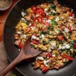 A skillet with Greek scrambled eggs, diced ham, cherry tomatoes, spinach, and crumbled feta cheese, with a wooden spoon resting inside.
