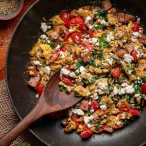 A skillet with Greek scrambled eggs, diced ham, cherry tomatoes, spinach, and crumbled feta cheese, with a wooden spoon resting inside.