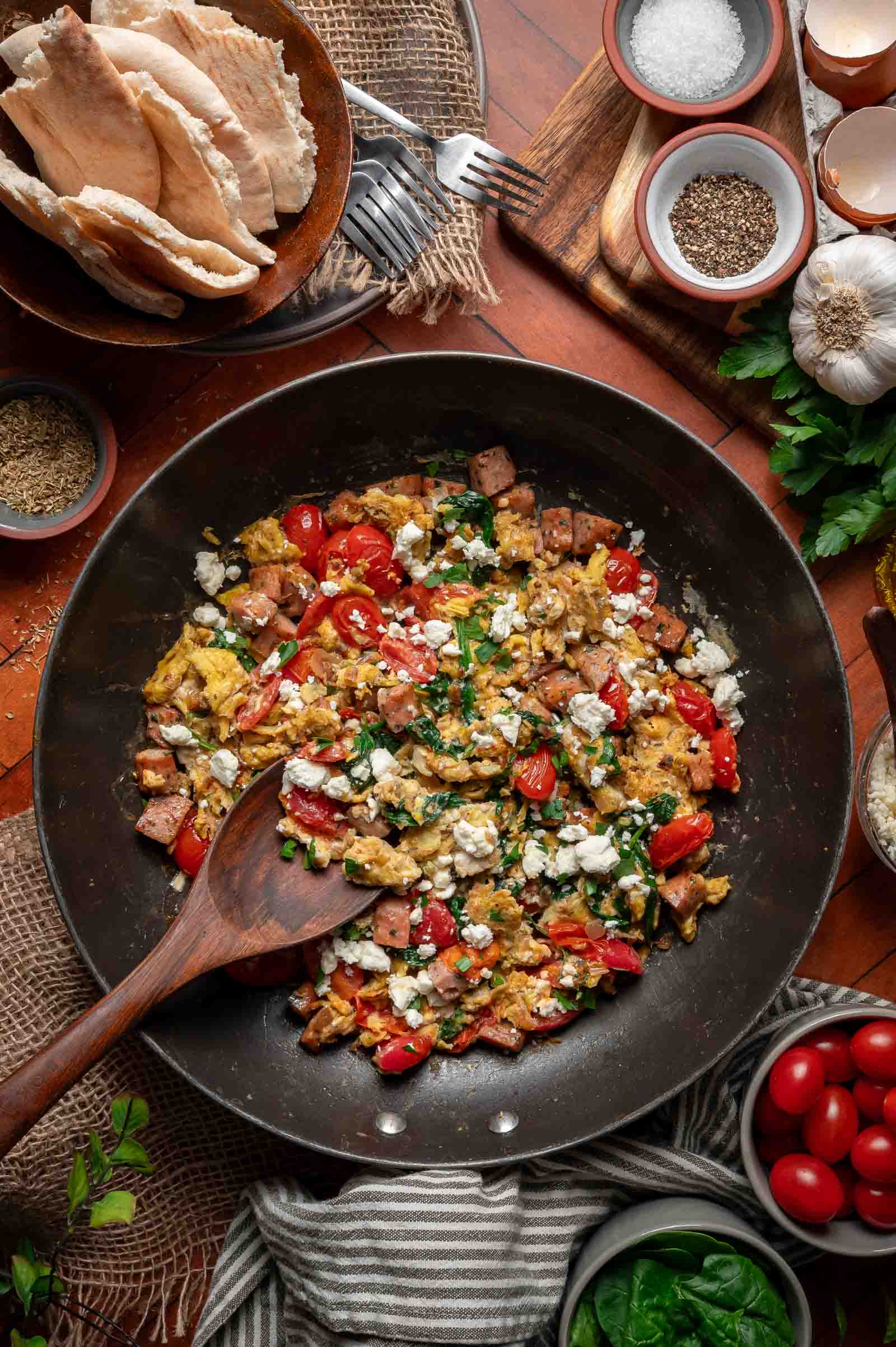A skillet with Greek scrambled eggs, diced ham, cherry tomatoes, spinach, and crumbled feta, surrounded by bowls of pita bread, spices, and fresh vegetables on a wooden table.