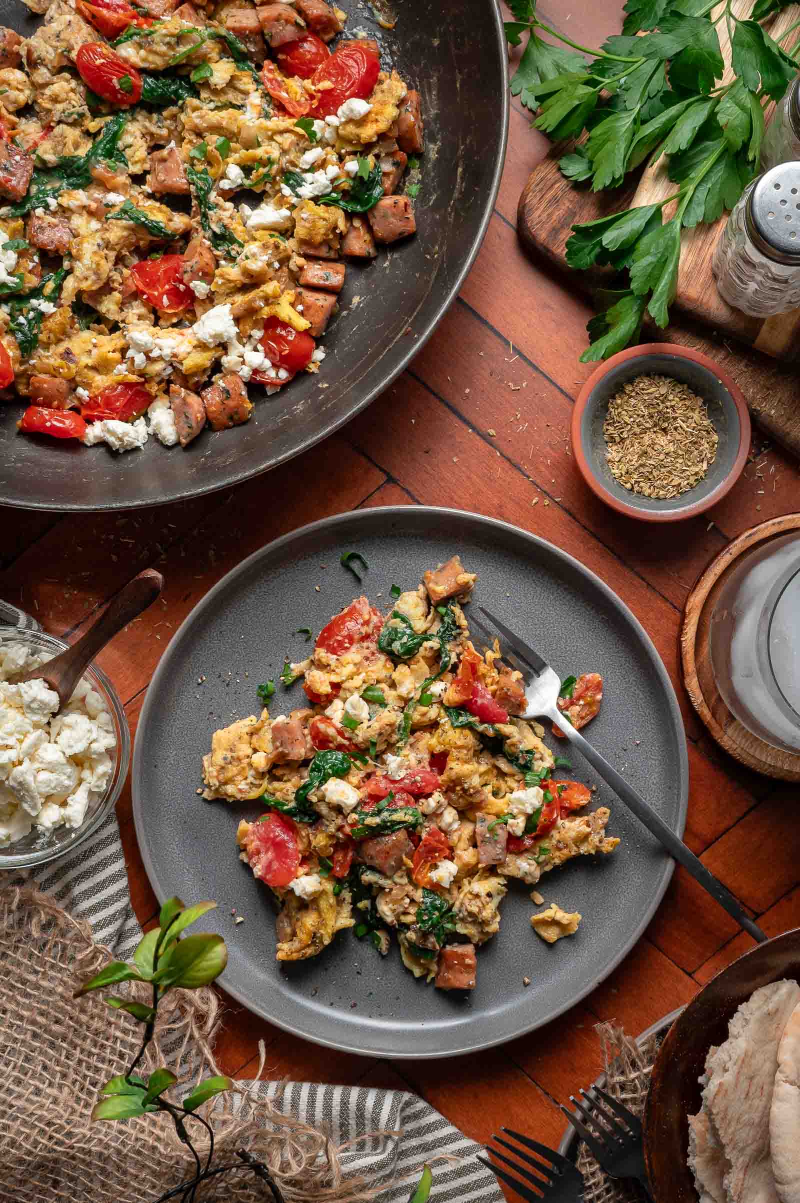Greek scrambled eggs with sausage, spinach, cherry tomatoes, and feta on a gray plate with a fork, surrounded by ingredients and a pan with more of the same dish.