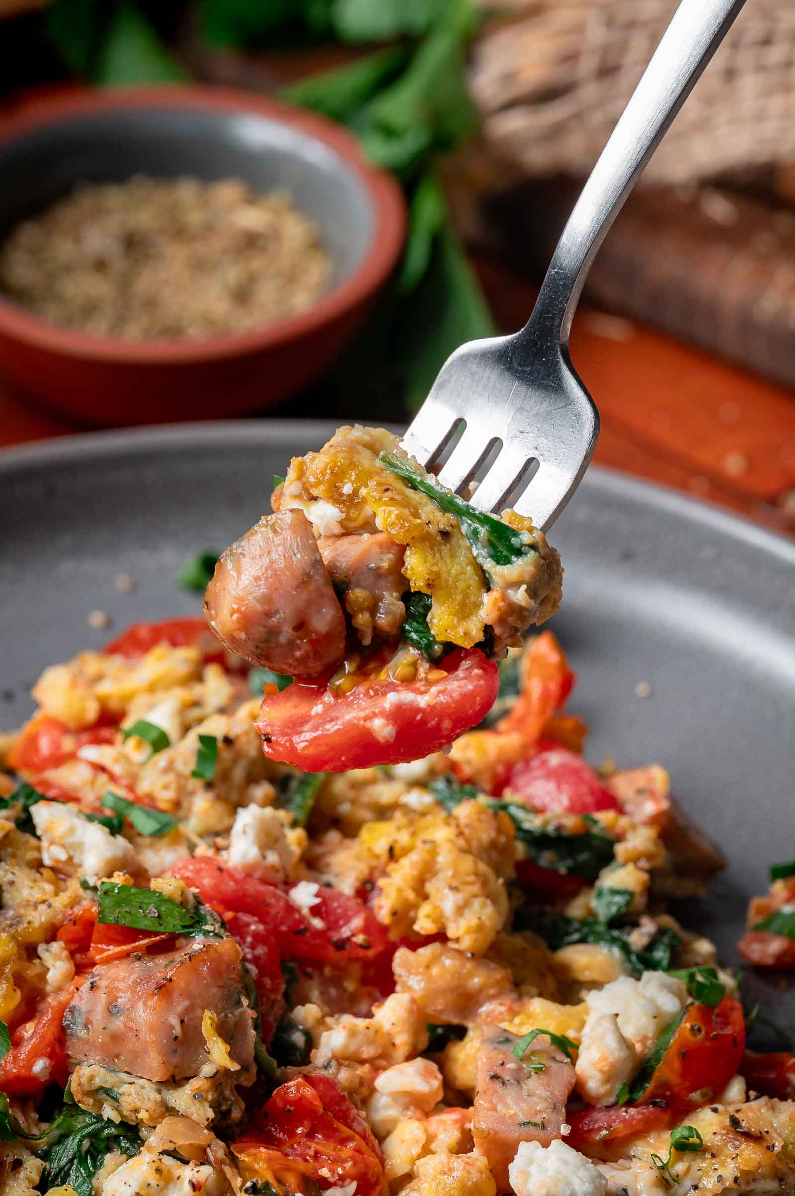 A fork holds a bite of Greek scrambled eggs mixed with sausage, tomatoes, spinach, and crumbled cheese over a plate of the same dish.