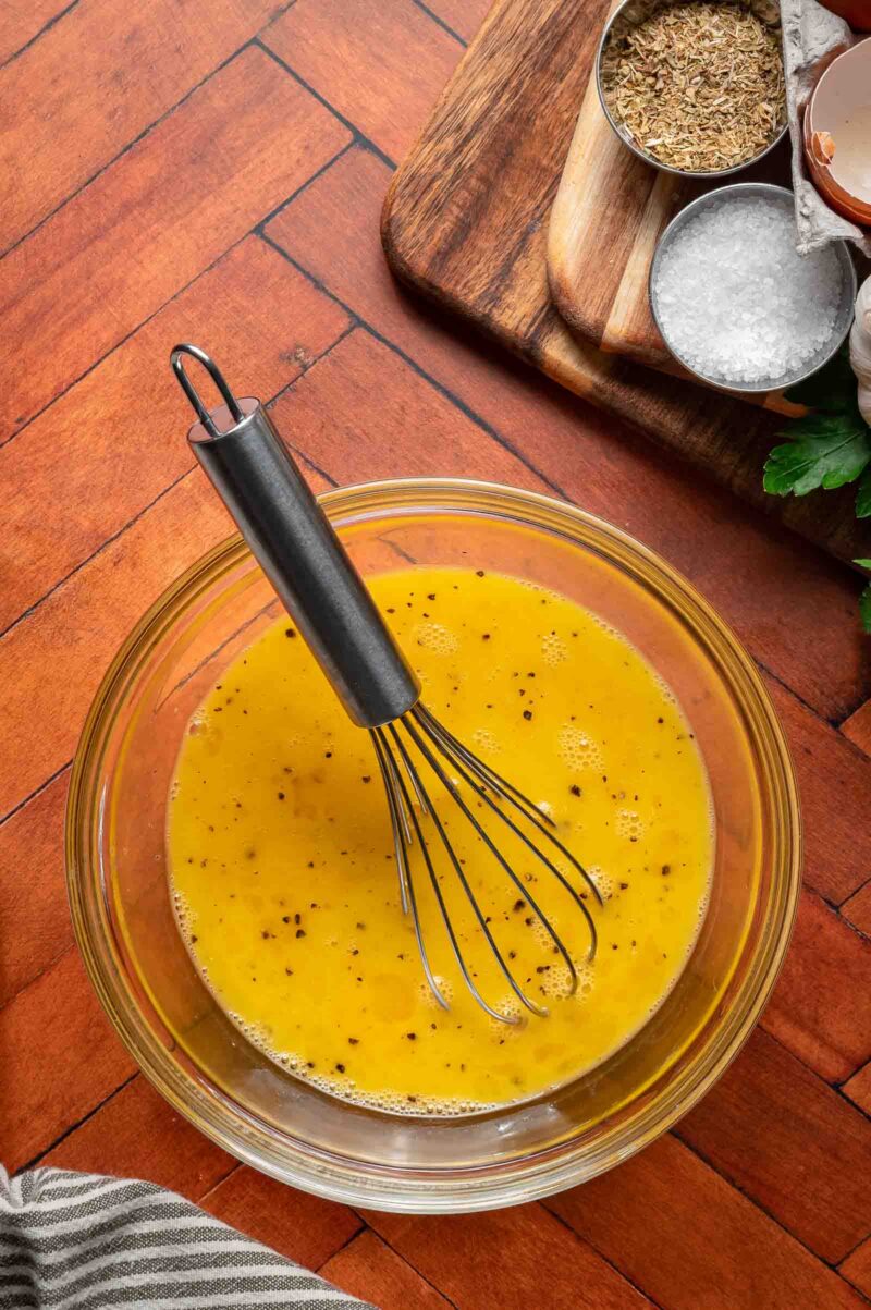 A glass bowl with whisked eggs and black pepper, a metal whisk inside. Nearby are bowls of seasoning and salt on a wooden board.