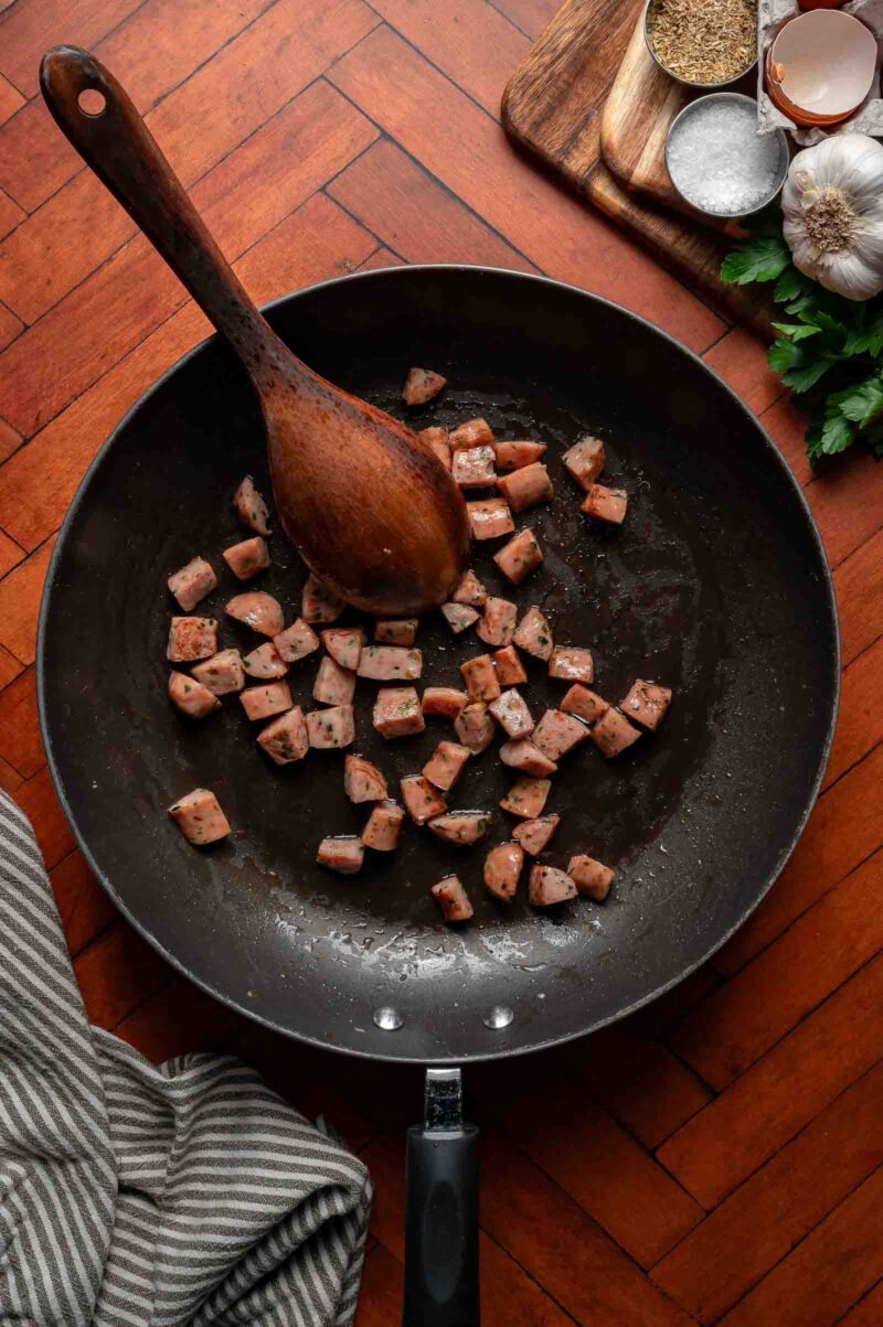 Diced sausage pieces are being cooked in a frying pan with a wooden spoon, seen from above. Spices, garlic, and herbs are arranged on a wooden board nearby.