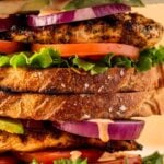 Close-up of a grilled chicken sandwich with lettuce, tomato, red onion, and multiple slices of toasted bread, stacked and served with potato chips. Text overlay reads "Grilled Chicken Sandwich.