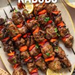 Overhead view of grilled lamb kabobs with red peppers and onions, garnished with herbs, served with lemon wedges and flatbread on a plate.
