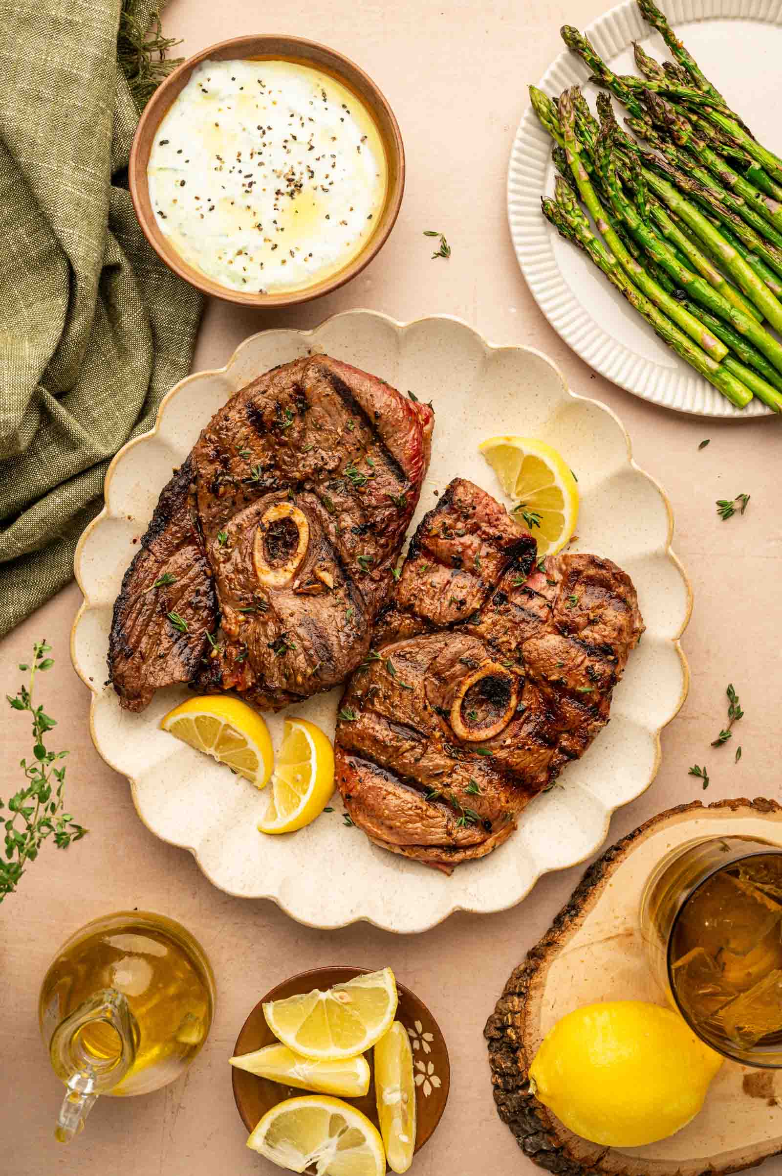Two grilled lamb steaks with bone on a plate with lemon wedges, surrounded by grilled asparagus, yogurt sauce, fresh herbs, and glasses of white wine on a tan table.