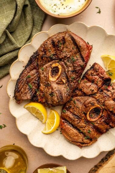 Two grilled lamb steaks with bone, garnished with herbs and served with lemon wedges on a scalloped white plate.
