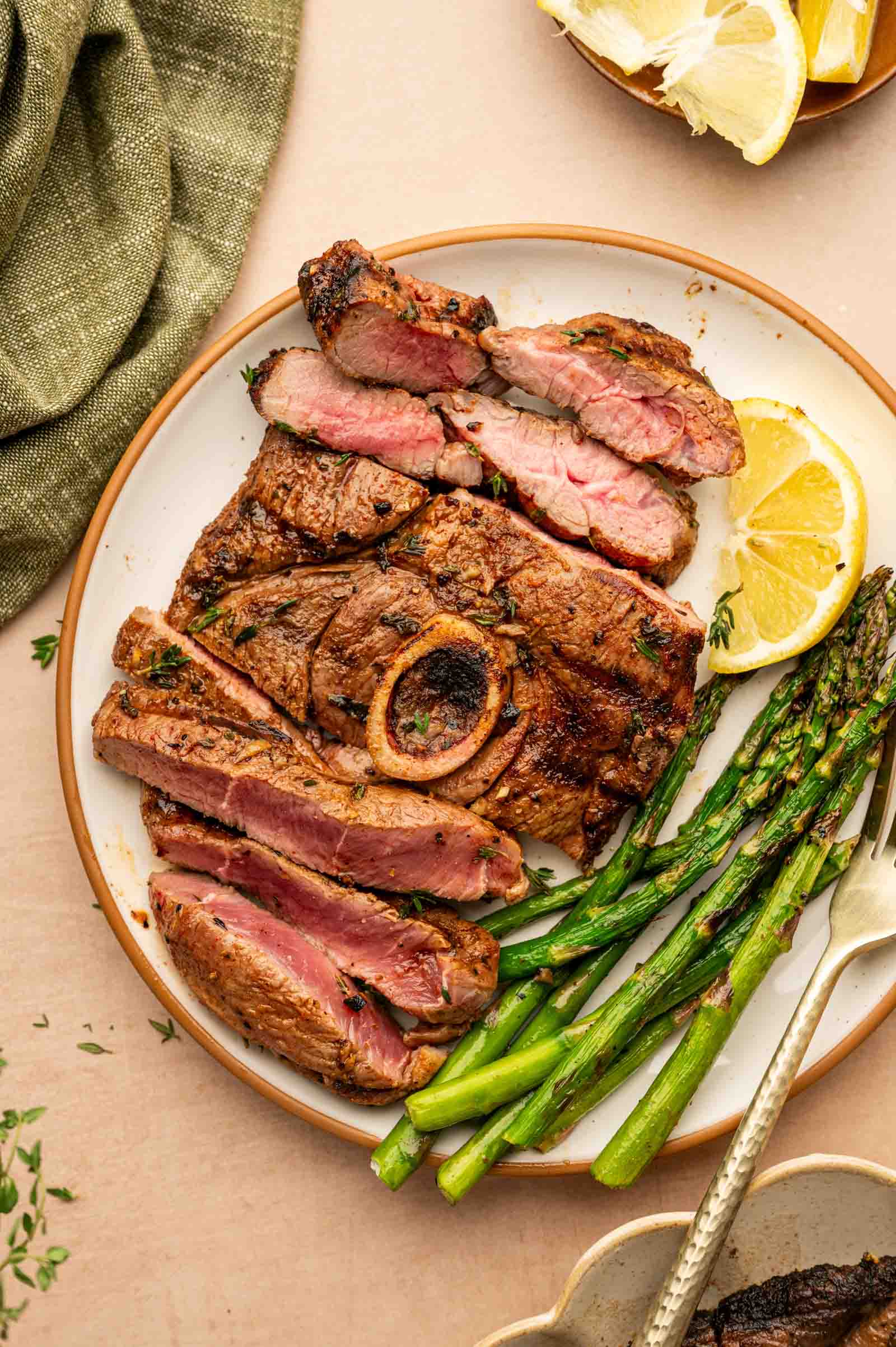 A plate with sliced cooked grilled lamb leg steak, a bone in the center, grilled asparagus, and a lemon wedge, with a fork on the side.