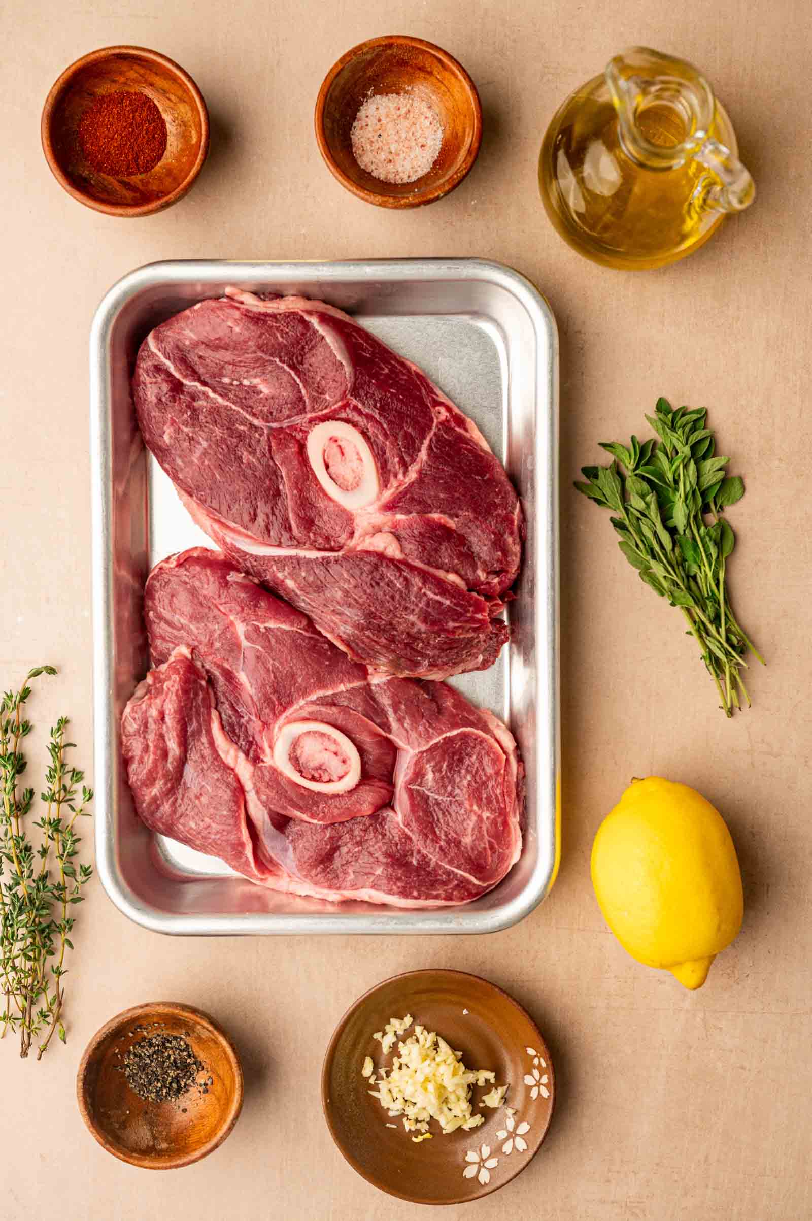 Two raw lamb leg steaks on a metal tray, surrounded by small bowls of oil, salt, pepper, spices, chopped garlic, fresh herbs, and a whole lemon on a beige surface.