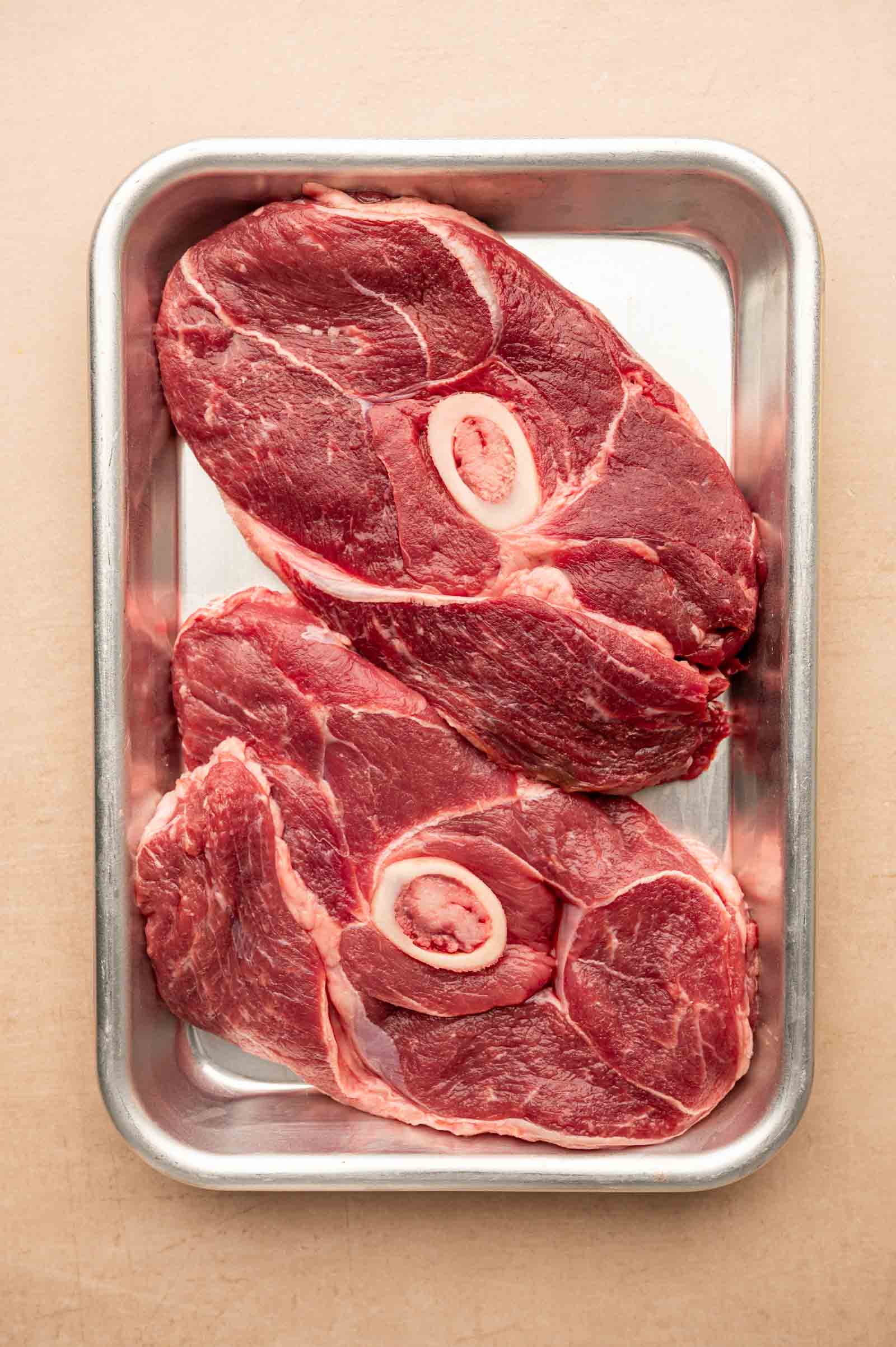 Two raw lamb leg steaks with marrow bone centers placed in a rectangular metal tray on a light-colored surface.