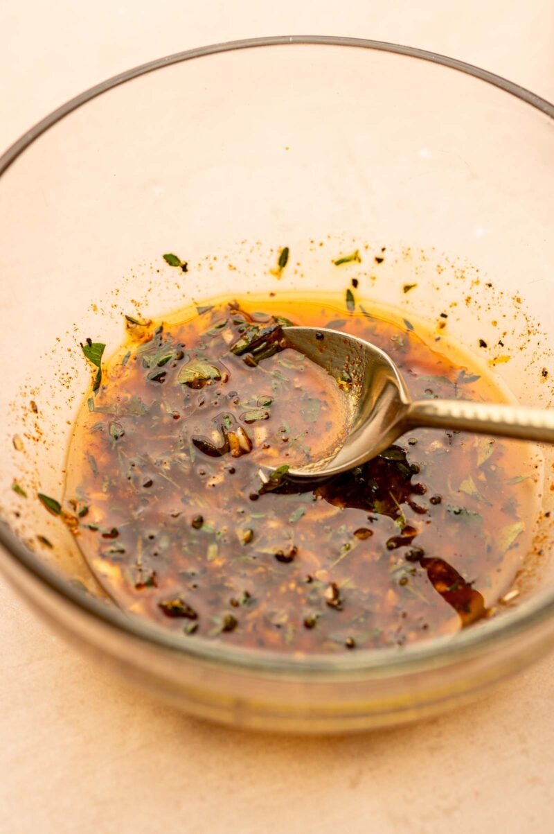 A glass bowl containing a mixed marinade with herbs and spices, and a metal spoon resting inside.