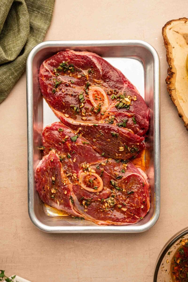Two raw lamb leg steaks marinated with herbs and spices are placed in a rectangular metal tray on a light-colored surface.
