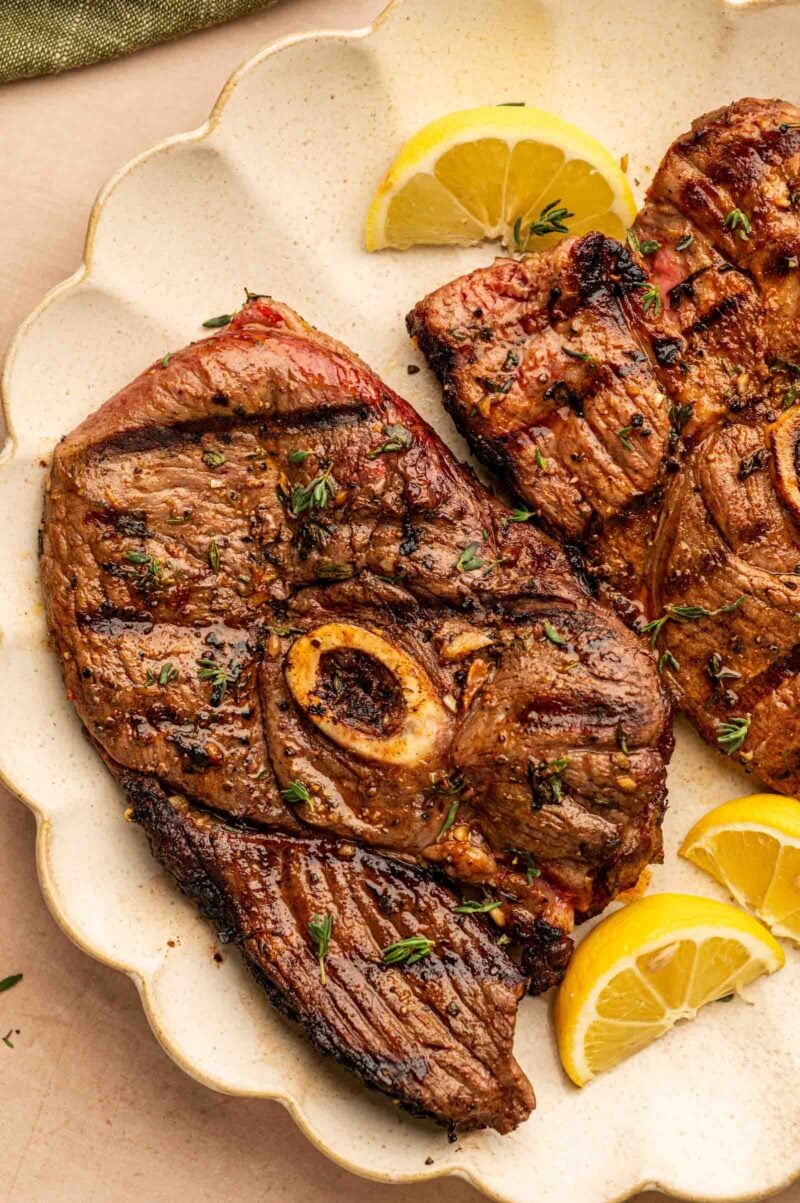 Grilled lamb leg steak with a bone served on a plate, garnished with fresh herbs and lemon wedges.