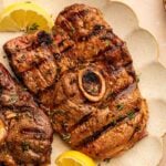 Grilled lamb leg steak with a bone in the center, garnished with fresh herbs and lemon wedges on a beige scalloped plate.