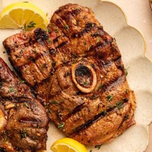 Grilled lamb leg steak with a bone in the center, garnished with fresh herbs and lemon wedges on a beige scalloped plate.