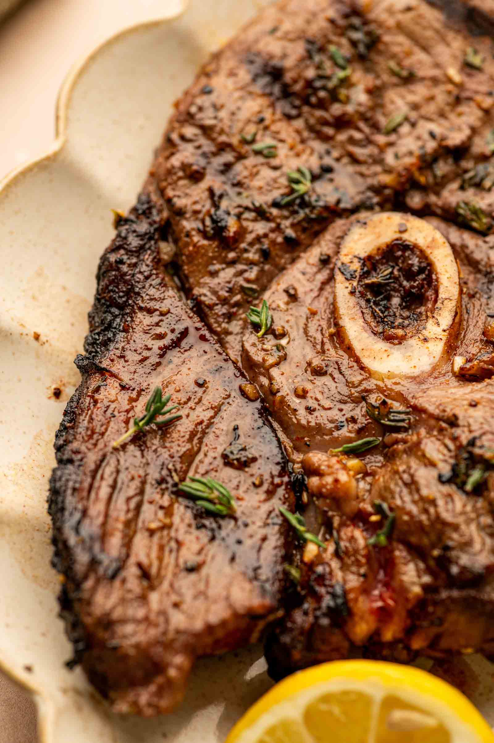 Close-up of a grilled lamb leg steak with a bone in the center, garnished with fresh herbs and accompanied by a slice of lemon on a beige plate.