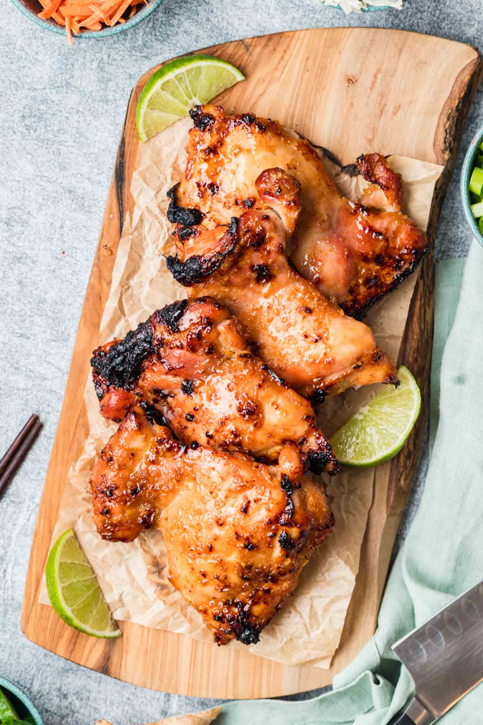 Cooked marinated lemongrass chicken thighs arranged on parchment paper atop a wooden board, garnished with lime wedges.