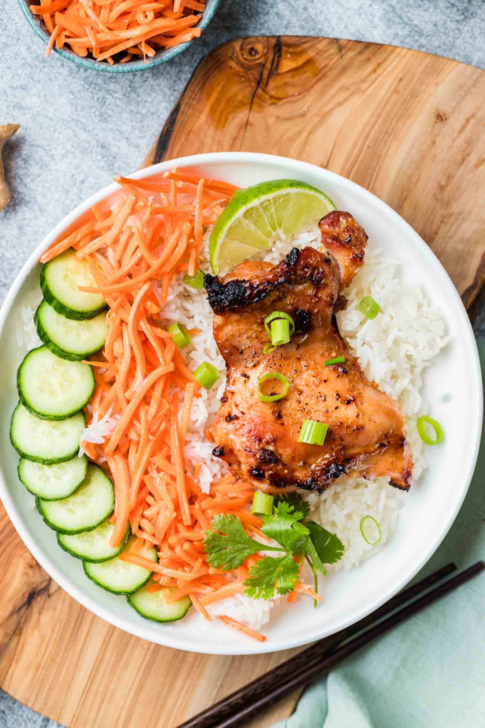 A bowl of white rice topped with lemongrass chicken, sliced cucumbers, shredded carrots, green onions, cilantro, and a lime wedge.