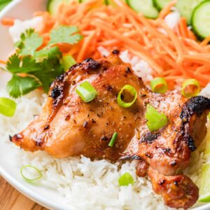Lemongrass chicken served on white rice with sliced cucumbers, shredded carrots, and cilantro, garnished with chopped green onions.