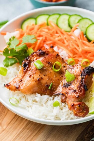Lemongrass chicken served on white rice with sliced cucumbers, shredded carrots, and cilantro, garnished with chopped green onions.