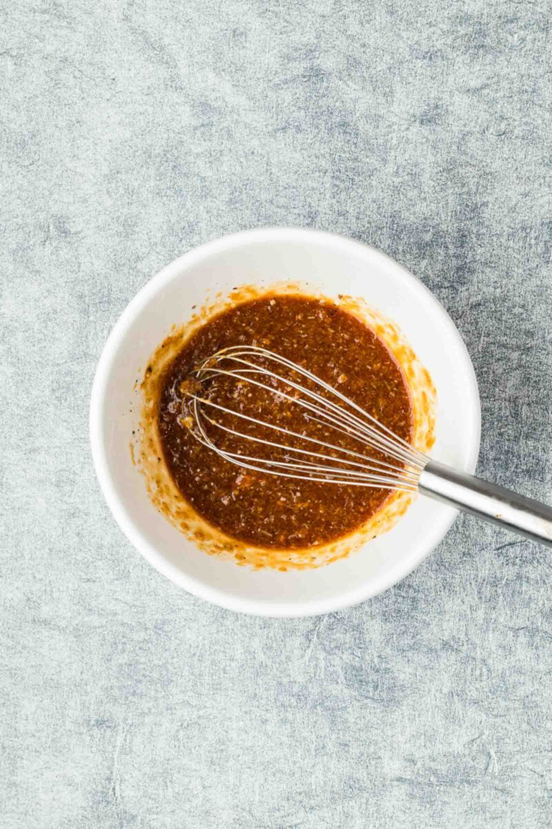 A metal whisk rests in a white bowl filled with a brown, mixed marinade on a textured gray surface.