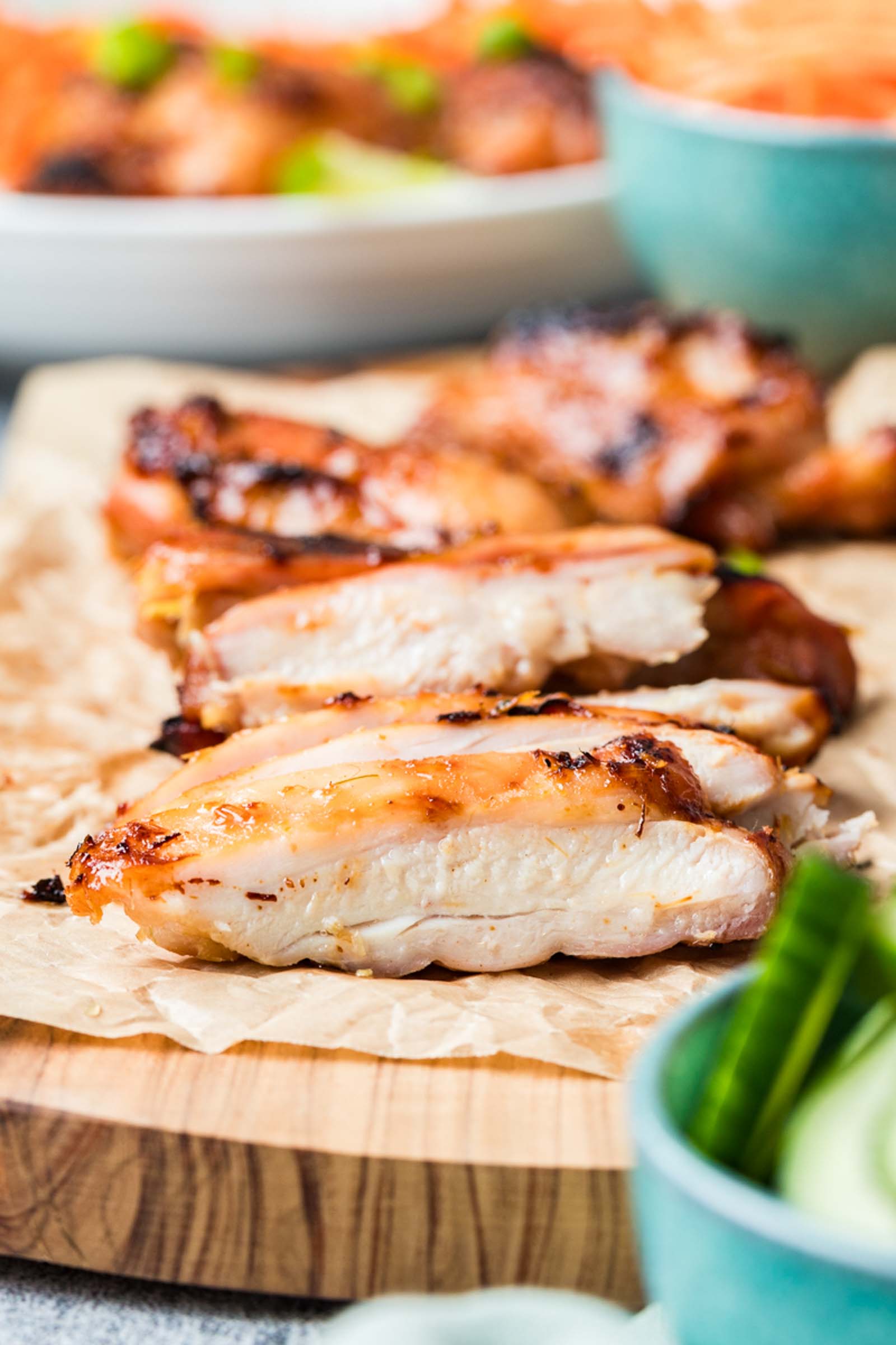 Sliced lemongrass chicken breast arranged on parchment paper atop a wooden board, with green vegetables and bowls in the background.