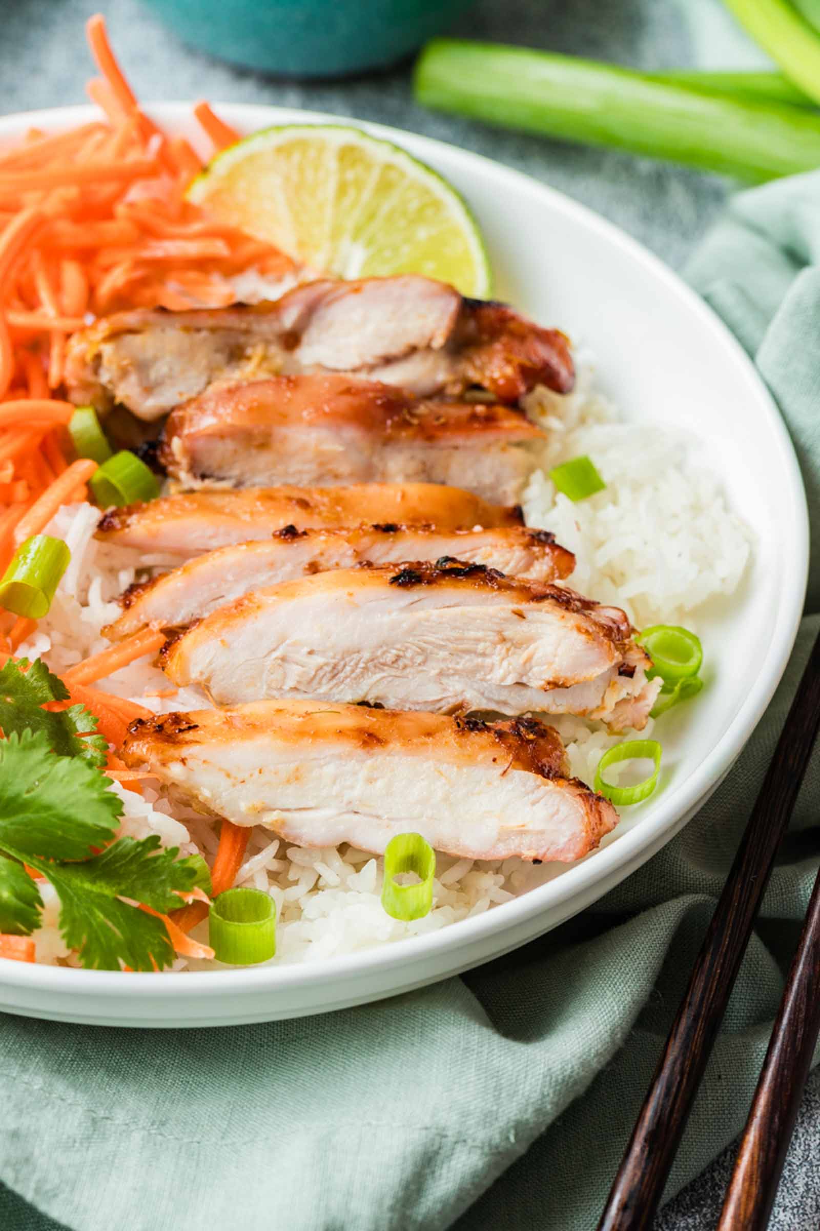 Sliced lemongrass chicken served over white rice with shredded carrots, green onions, cilantro, and a lime wedge on a white plate.