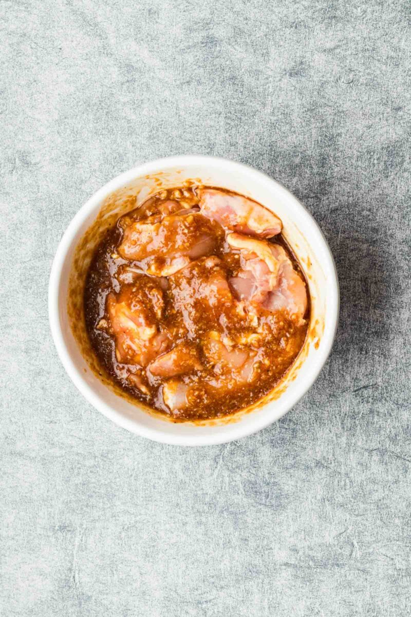 Raw chicken pieces marinating in a lemongrass sauce in a white bowl, placed on a textured gray surface.