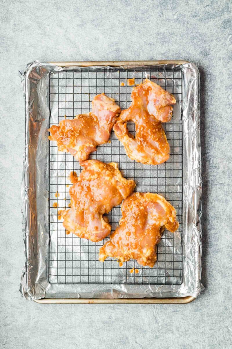 Four pieces of marinated raw chicken on a wire rack over a foil-lined baking sheet.